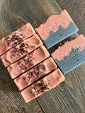 Image shows a group of black and pink soaps with rose petals and black sea salts on top
