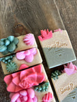 Image shows a group of green and tan soaps topped with colorful succulent soaps